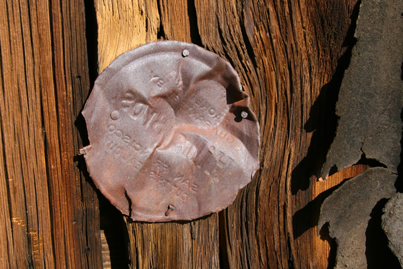 The top of a Hills Bros. coffee tin repurposed to cover a knothole in the wood siding.